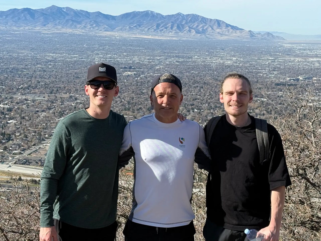 A father and his two sons standing together outdoors with mountains behind them.
